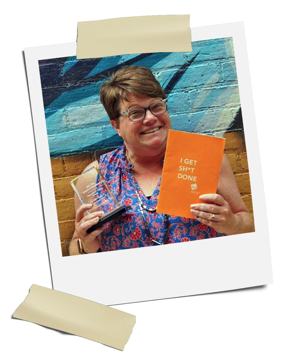 A Polaroid photo of a woman, Crissy, with short hair and glasses holding an orange notebook that says "I Get Sh*t Done" in one hand, and an award in the other. She is wearing a blue and red print, sleaveless top and is standing in front of a brick wall with graffiti on it.